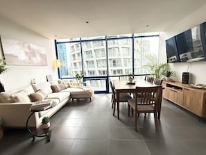 En moderne stue med sofa, spisebord, stole, store vinduer med byudsigt og et fladskærms-tv monteret på væggen.