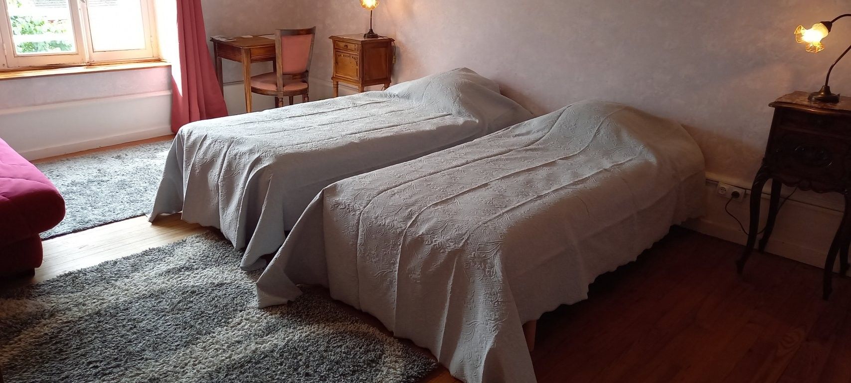 Een kamer met twee eenpersoonsbedden, een klein houten tafeltje, een stoel, een rode bank en een raam met gordijnen.