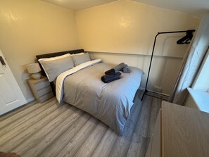 Een netjes opgemaakt bed met hoofdbord, een nachtkastje met een lamp, en een kledingkast met daarin hangende kleding.