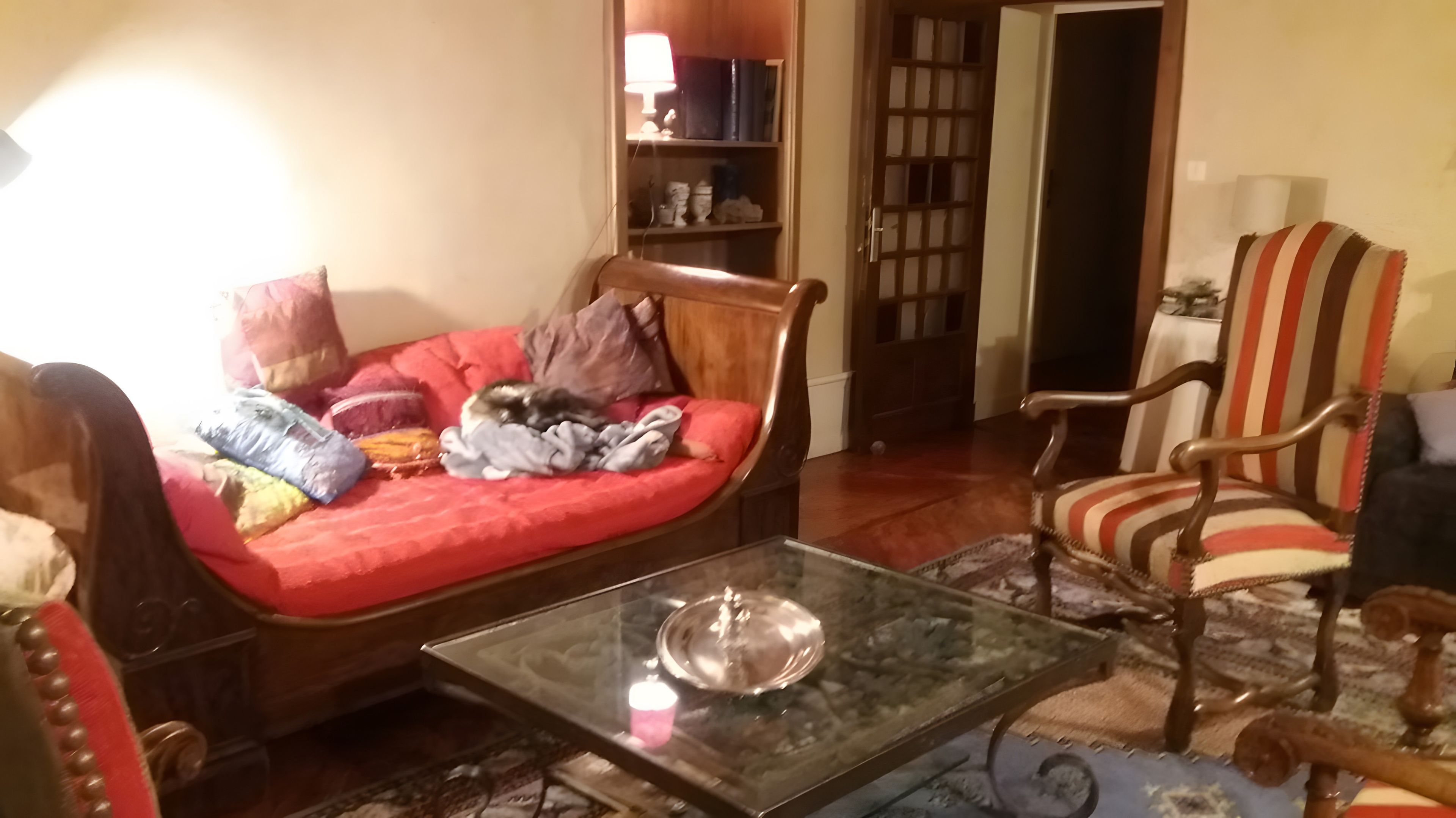 Een woonkamer met een rode bank, een gestreepte fauteuil, een glazen salontafel en een boekenkast.