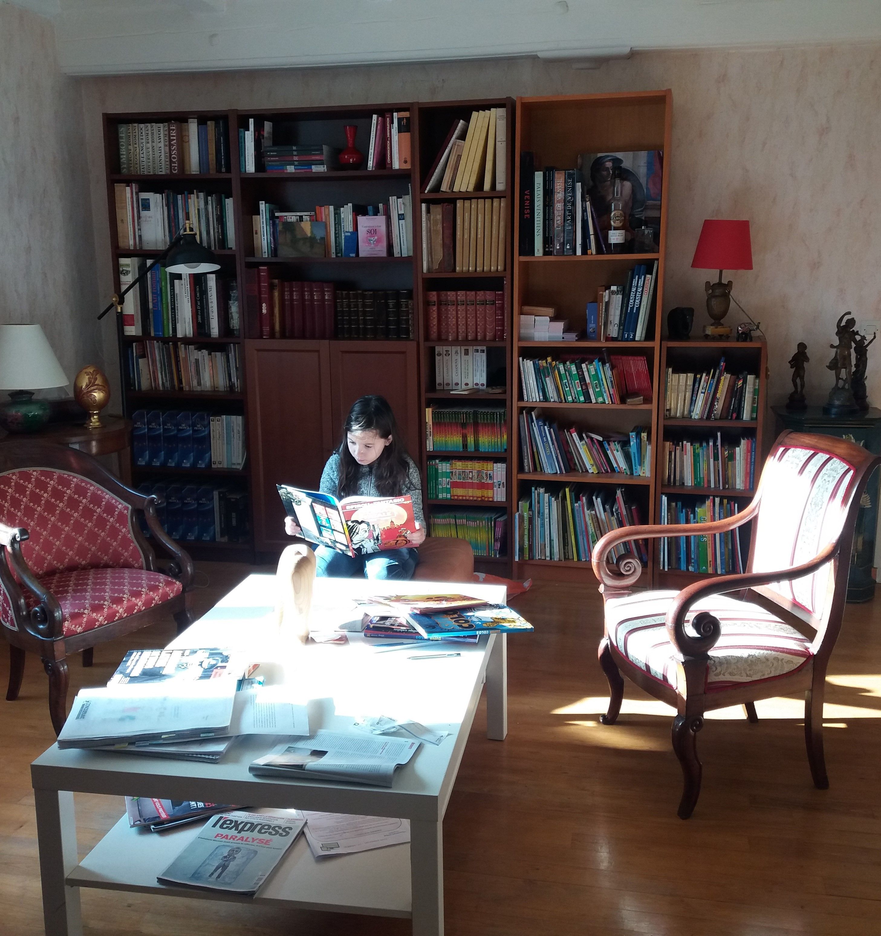 Een kamer met daarin een persoon die een boek leest, omringd door boekenplanken vol boeken, een tafel met kranten en twee stoelen.
