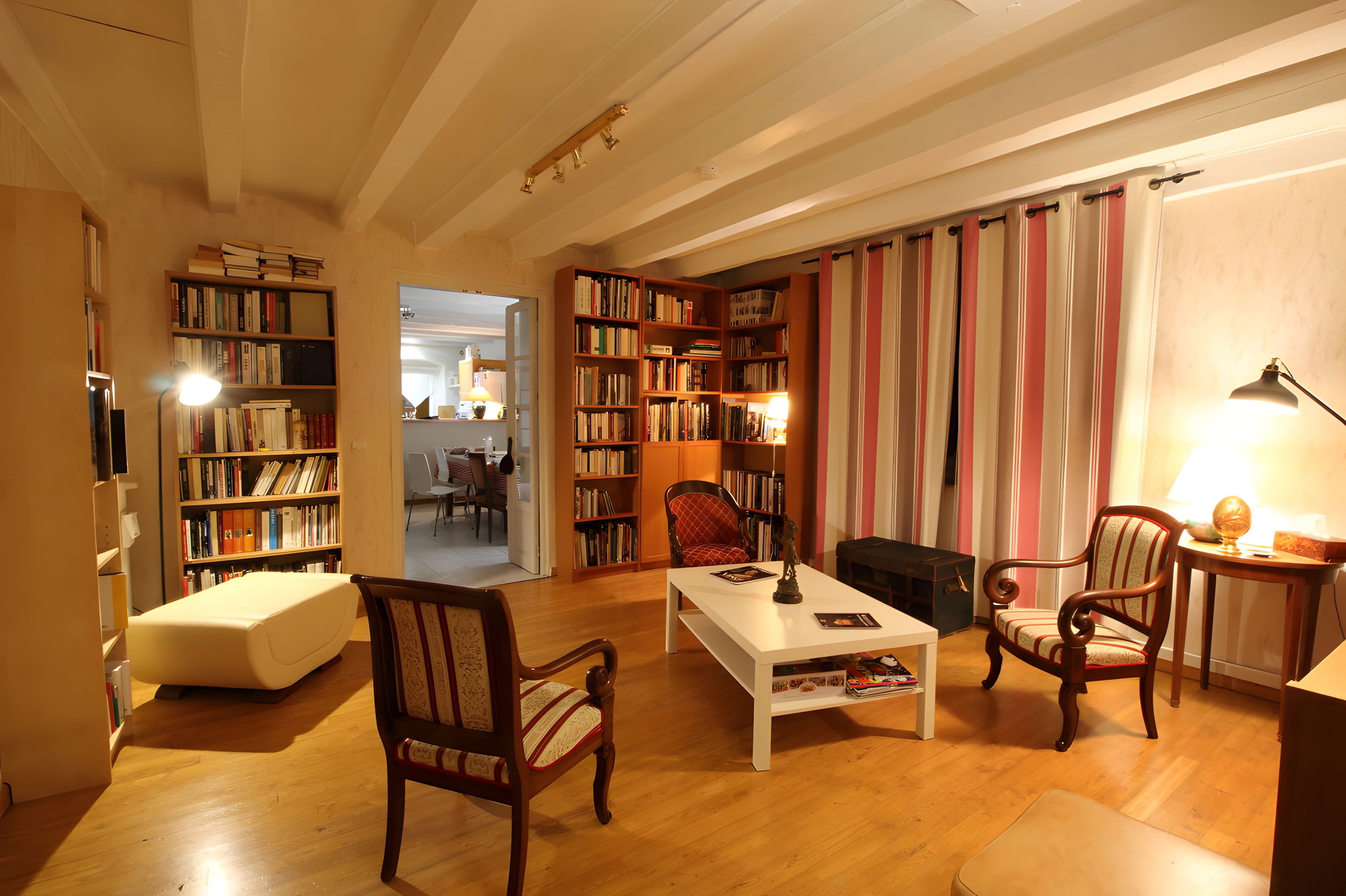 Een gezellige woonkamer met een open haard, boekenkasten, een eethoek en een doorkijk naar een andere kamer.