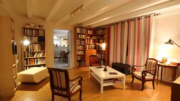 Een gezellige woonkamer met een open haard, boekenkasten, een eethoek en een doorkijk naar een andere kamer.