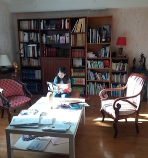 Une pièce où une personne est assise, en train de lire un livre, entourée d’étagères remplies de livres, d’une table avec des journaux et de deux chaises.