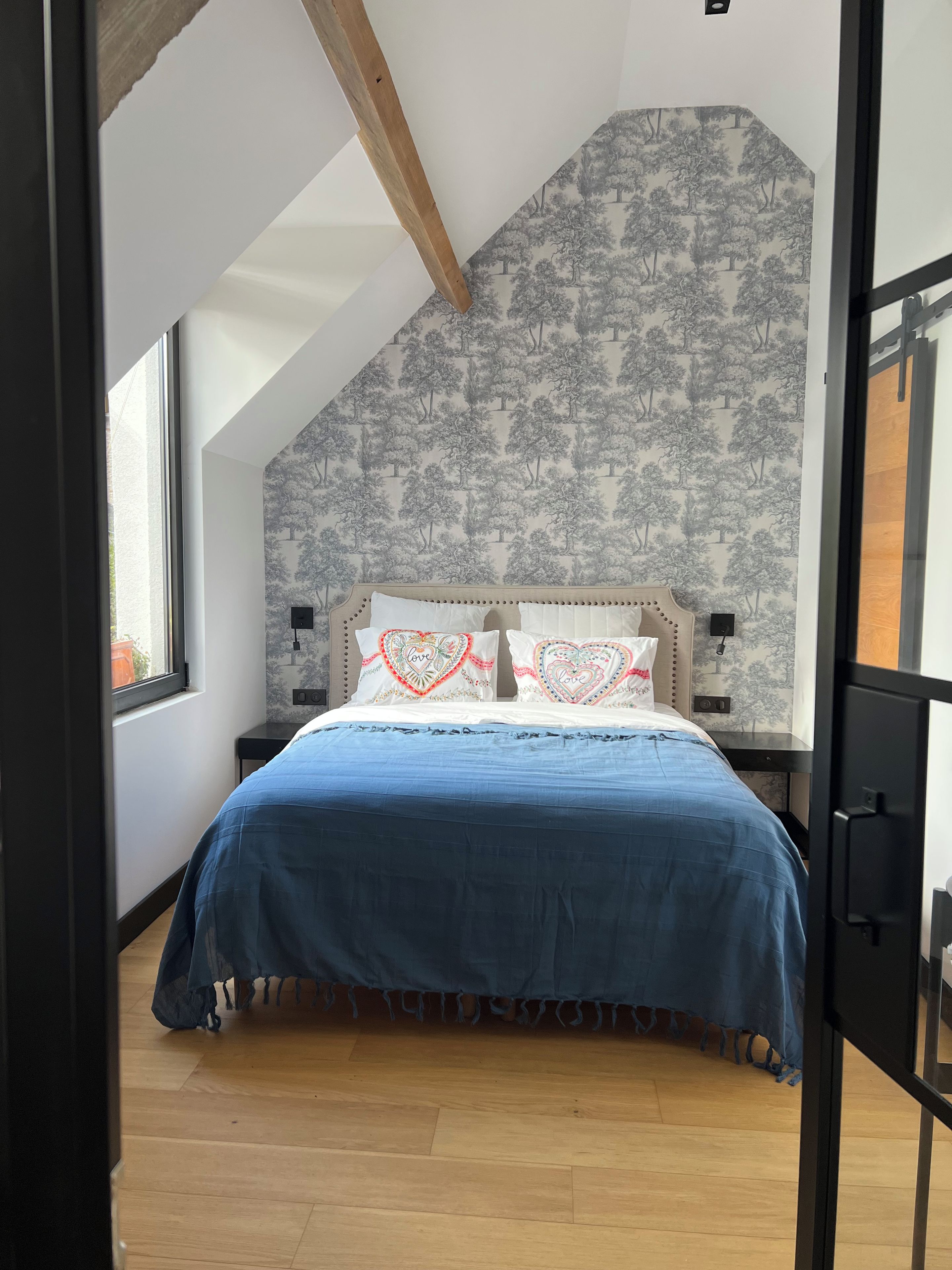 Une chambre avec un plafond en pente, un lit recouvert d’une courtepointe bleue, deux coussins à motifs de cœur et un mur décoré d’un motif d’arbre.