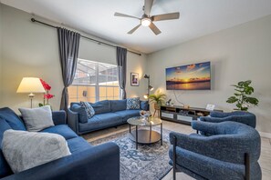 A modern living room with a blue sofa set, a coffee table, a floor lamp, and a ceiling fan. - 3BR Townhome with Private Pool at Paradise Palms Near Disney (Kissimmee)