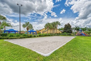 A sandy volleyball court surrounded by green grass, with palm trees and a playground in the background. - 3BR Townhome with Private Pool at Paradise Palms Near Disney (Kissimmee)