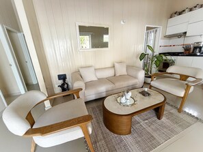 A living room with a beige sofa, two armchairs, a coffee table, and a kitchen area with cabinets and a plant.