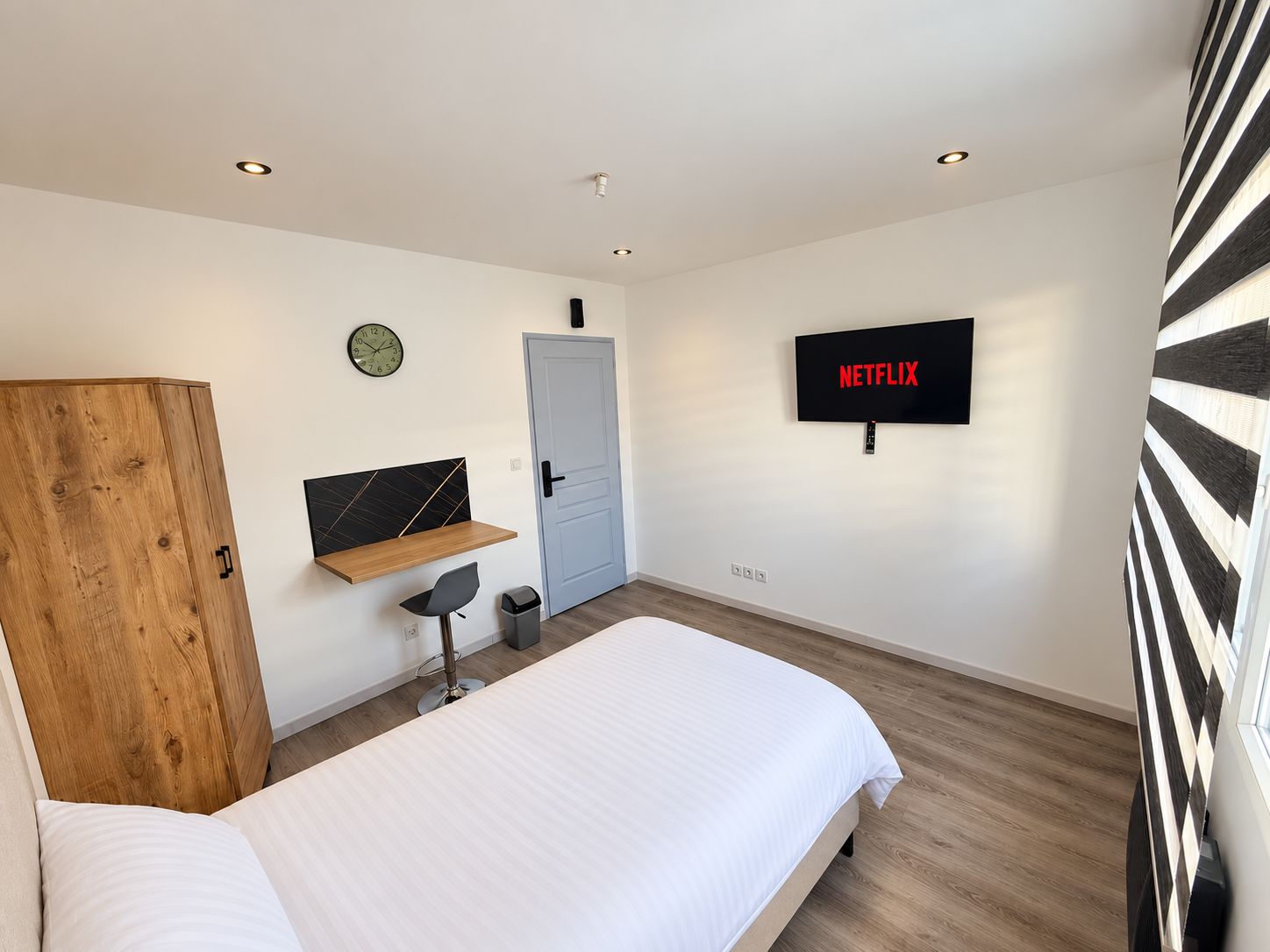 Et moderne soveværelse med seng, træskab, skrivebord med stol, vægmonteret TV og en dør.