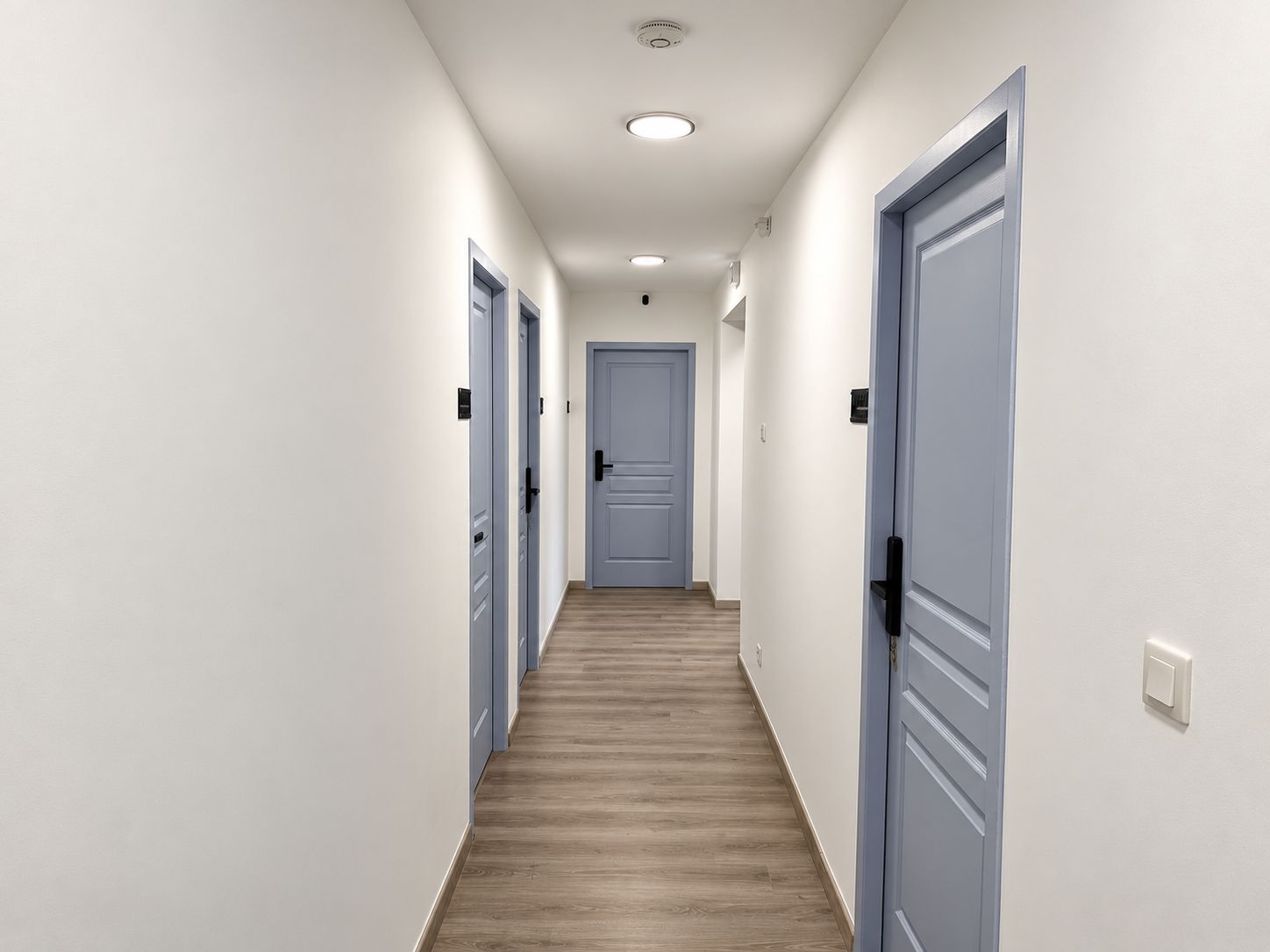 A hallway with light-colored walls, wooden flooring, and blue doors.
