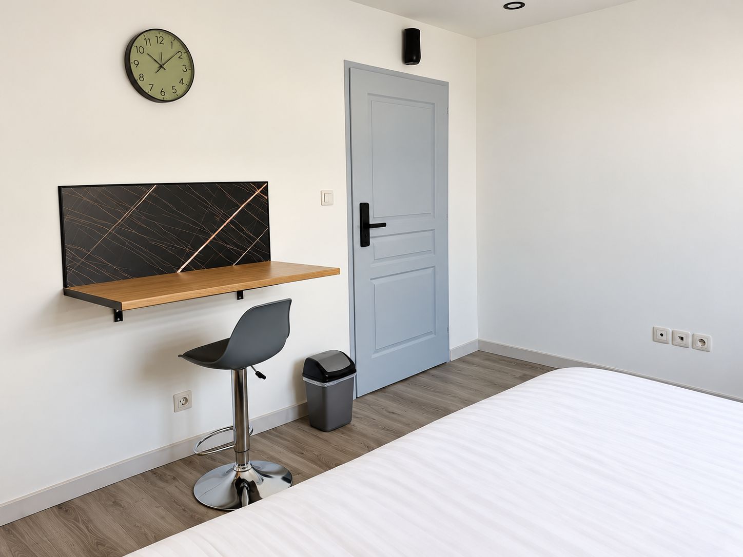 Een slaapkamer met een bed, een bureau met stoel, een klok en een deur.