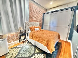 A bedroom with a brick wall, a bed with an orange bedspread, a nightstand with a lamp, and a wardrobe. - Soothing 1-bedroom apartment with AC in Birmingham (Birmingham)