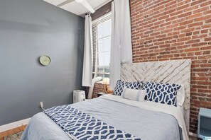 A hotel room with a brick wall, a bed with patterned bedding, a clock on the wall, and a window with curtains. - Soothing 1-bedroom apartment with AC in Birmingham (Birmingham)