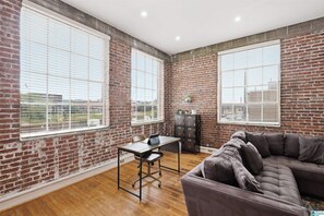A room with brick walls, large windows, a wooden floor, a desk with a chair, and a sectional sofa. - Soothing 1-bedroom apartment with AC in Birmingham (Birmingham)