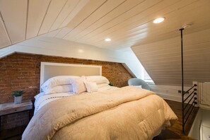 A bedroom with a sloped ceiling, a brick wall, a bed with patterned bedding, a nightstand with a plant, and a chair. - The Sycamore Downtown Modern Home (Lancaster)
