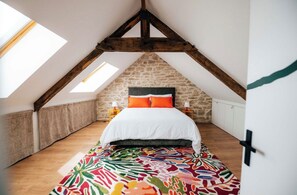 A bedroom with a sloped ceiling, a bed with white bedding and orange pillows, a colorful rug, and a stone wall. - Exceptional thatched cottage, Nordic bath, and wood-burning fire (Ploemeur)
