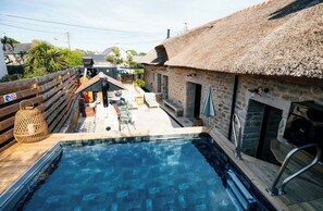 A swimming pool with a wooden deck, a thatched-roof building, and outdoor furniture. - Exceptional thatched cottage, Nordic bath, and wood-burning fire (Ploemeur)