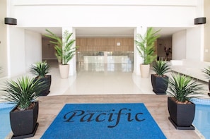 Eine Hotelhalle mit einer Rezeption, Blumentöpfen und einem blauen Teppich mit dem Aufdruck 'Pacific'.