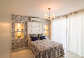 A bedroom with a bed, a chandelier, wall-mounted lights, and a window with curtains. - Luxury Beachfront Apartment in Arraial do Cabo – Sea Views, Pools & BBQ (Cabo Frio)