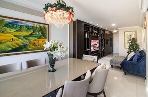 A dining area with a long table, chairs, a chandelier, a bookshelf, and a television. - Luxury Beachfront Apartment in Arraial do Cabo – Sea Views, Pools & BBQ (Cabo Frio)