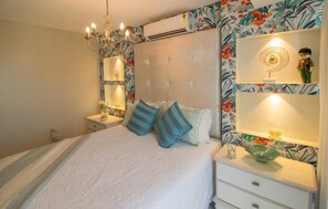 A bedroom with a bed, nightstands, a chandelier, and a wall-mounted shelf with decorative items. - Luxury Beachfront Apartment in Arraial do Cabo – Sea Views, Pools & BBQ (Cabo Frio)