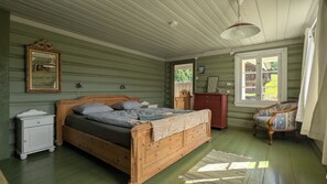 A room with a wooden bed, a nightstand, a dresser, a chair, and a window with a view of greenery. - Historic Norwegian farmhouse in Telemark (Telemark)