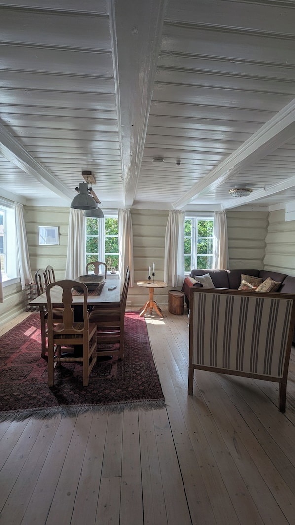 A spacious loft with a high ceiling, exposed wooden beams, a dining area with a table and chairs, a sofa, and large windows offering views of greenery.