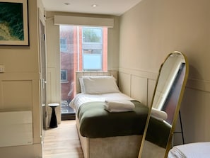Ett hotellrum med en säng, en liten pall, en stor spegel och ett fönster med utsikt över byggnader.