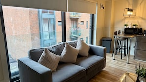 A modern living room with a grey sofa, a large window, and a kitchen area with a microwave and bar stools. - Stylish 1-Bed Apartment in the heart of Manchester (Unknown)