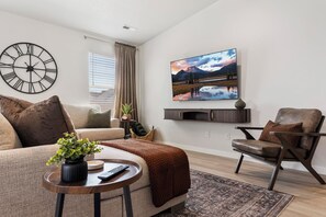 En moderne stue med et stort fladskærms-TV på væggen, en behagelig sofa, et sofabord med en plante og en stilfuld lænestol.