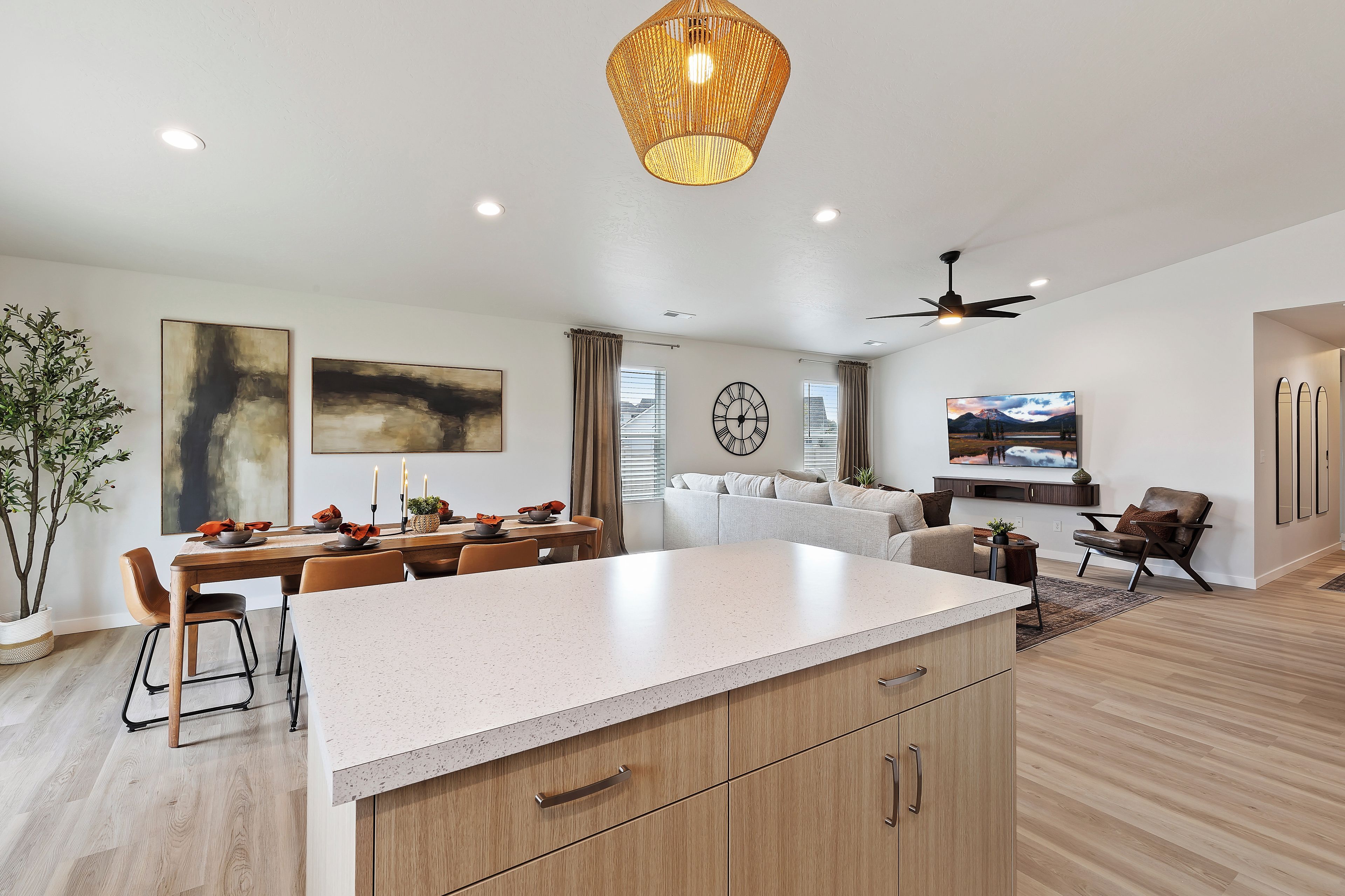Een moderne keuken met een centraal eiland, een eetgedeelte met stoelen en een gedekte tafel, een woonkamer met een bank en een televisie, en een plafondventilator.