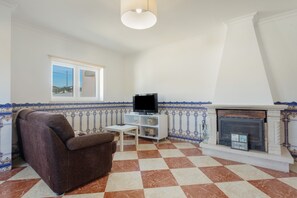 Un salon avec un sol à carreaux, un fauteuil marron, un téléviseur posé sur un meuble blanc, une cheminée ornée de carreaux décoratifs et une fenêtre donnant sur le ciel.