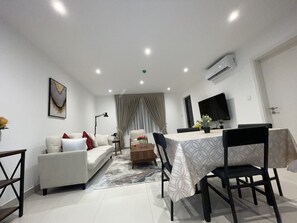 A modern living room with a sofa, a dining table with chairs, a TV mounted on the wall, and an air conditioning unit. - Luxurious 1-bedroom apartment Airport Residential (Accra)