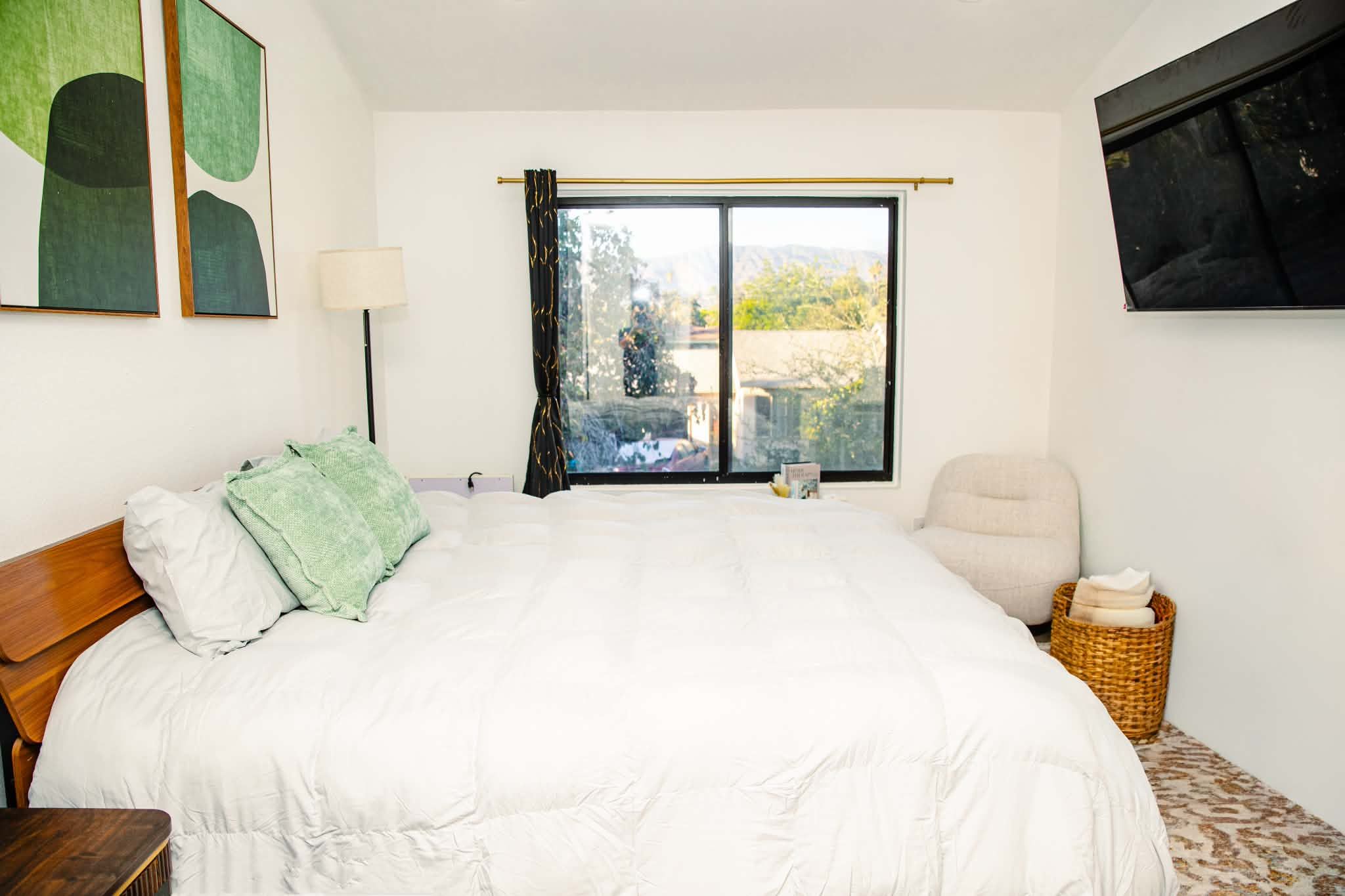 A bedroom with a large bed, a chair, a window with curtains, and a flat-screen TV mounted on the wall.