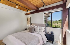 A bedroom with a large bed, a nightstand, a lamp, and a window with a view of the outdoors. - Hillview Ranch Retreat A/C Views 16+ guests (Fallbrook)