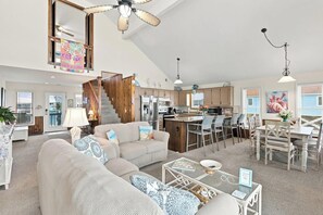A spacious living area with a high ceiling, a kitchen with modern appliances, a dining area with a bar, and a staircase leading to an upper level. - A Little Piece of Heaven | 4BR w/ Oceanfront Decks, Direct Beach Access & Views (Ocean Isle Beach)