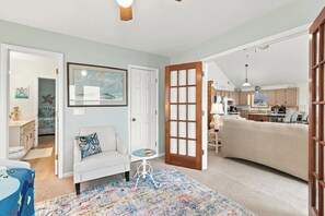 Une pièce avec un canapé blanc, une petite table ronde, un tapis coloré, une porte avec des panneaux de verre et une cuisine visible à travers une porte ouverte.
