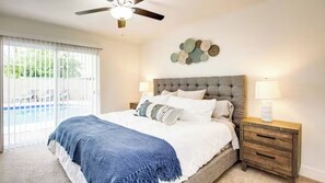 Ein Schlafzimmer mit einem großen Bett, einem Deckenventilator, einem Nachttisch mit Lampe und Blick auf einen Pool durch das Fenster.