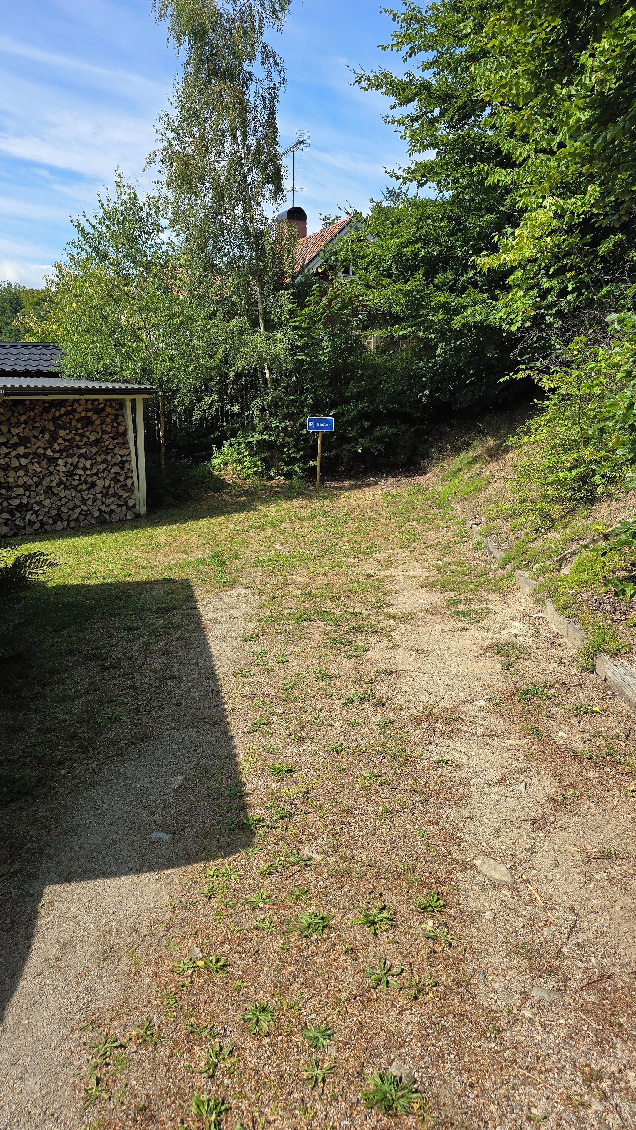 Ein Kiesweg führt zu einem Gebäude mit einem Holzstapel und einem blauen Parkschild.