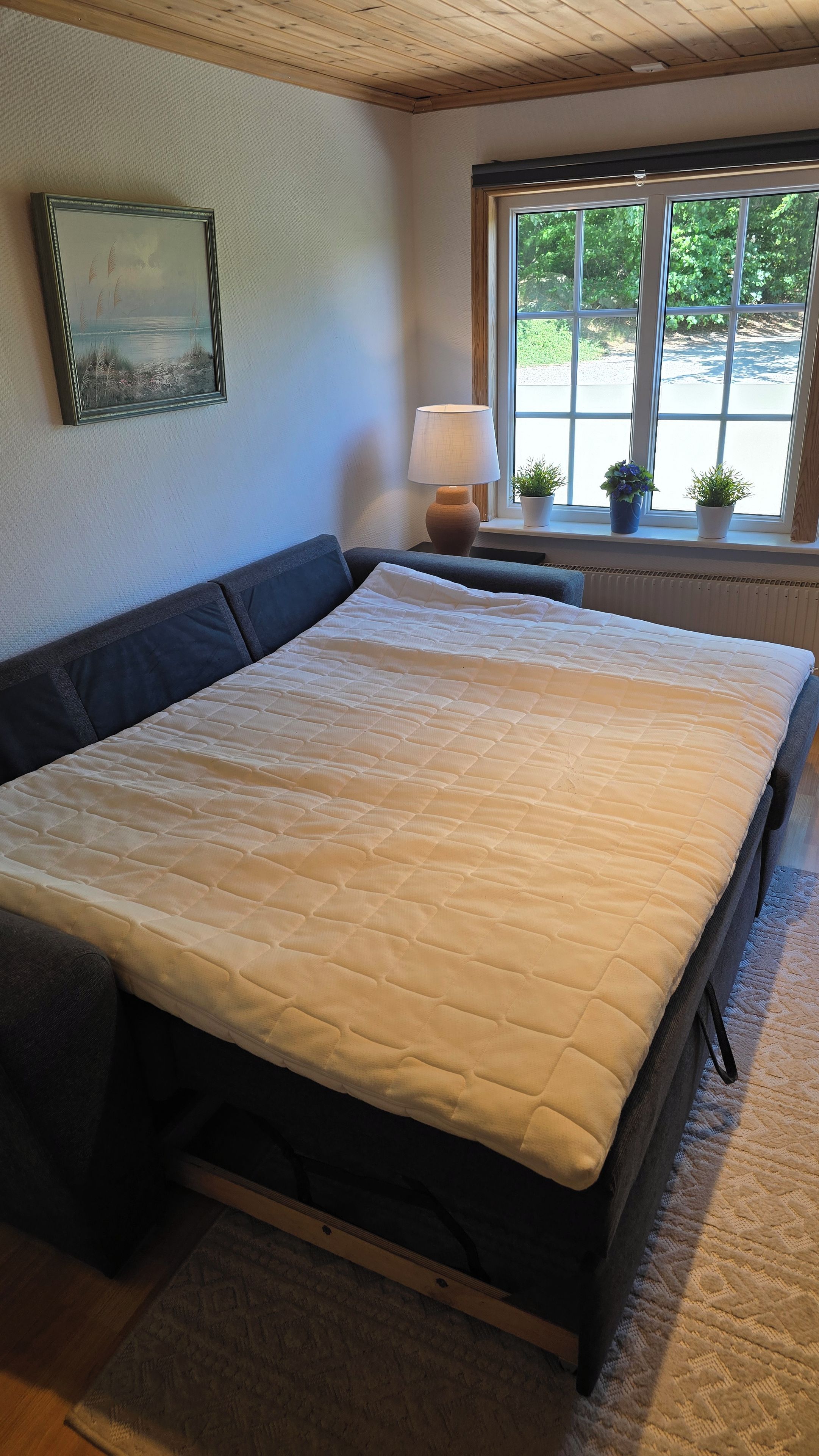 Ein Schlafzimmer mit Bett, Nachttisch, Lampe, Fenster mit Blick auf Grünflächen und einem gerahmten Bild an der Wand.