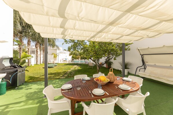 Un patio exterior con una mesa redonda de madera, sillas blancas, una parrilla y una zona cubierta.