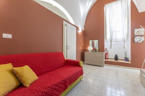 Quarto com sofá vermelho, cómoda de madeira, extintor de incêndio e janela com cortinas.
