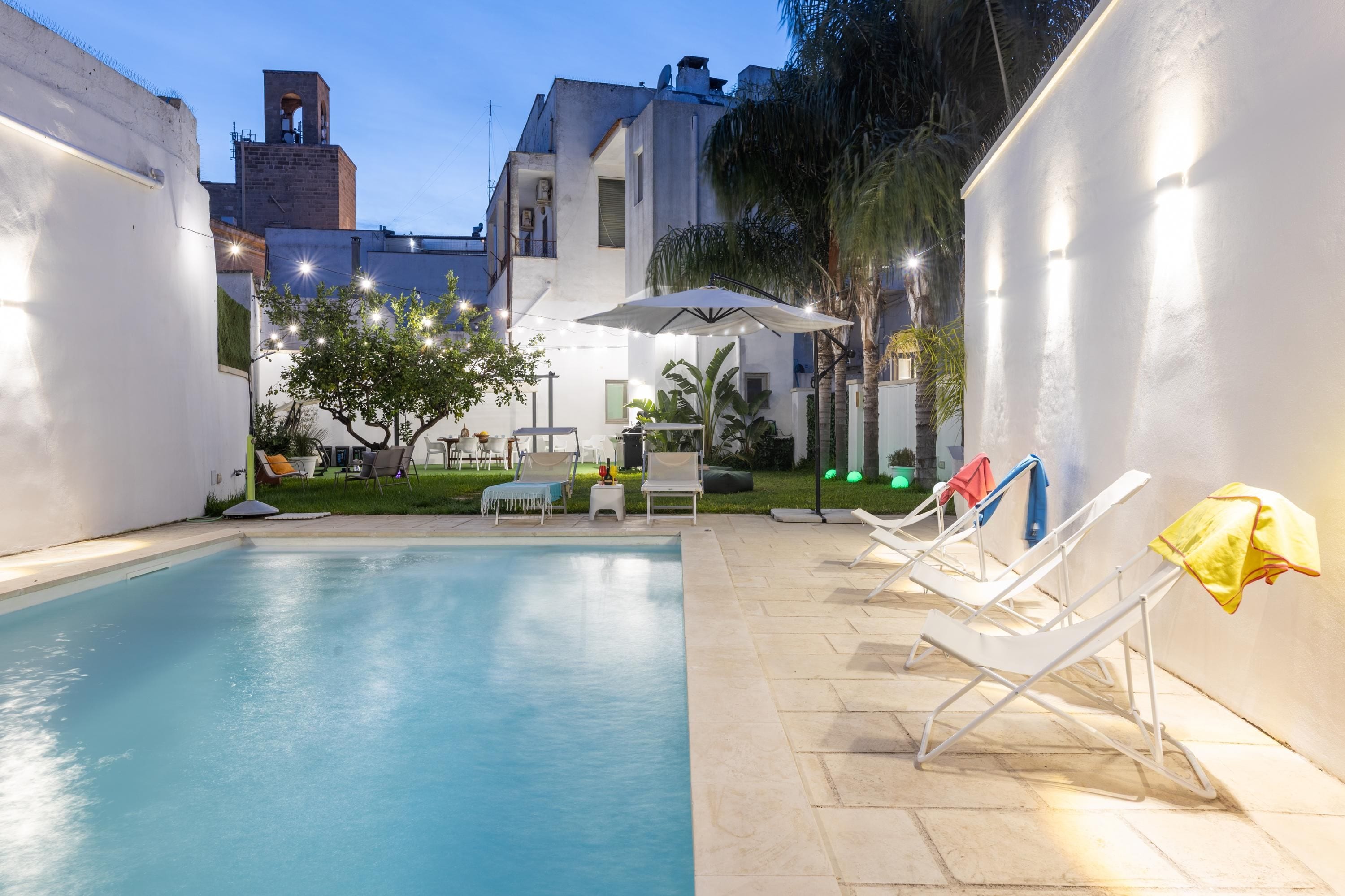 Area piscina esterna con sedie a sdraio e un tavolo, circondata da un muro bianco e un edificio con un campanile.