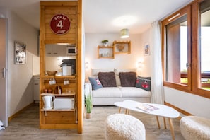 A cozy room with a sofa, a table, and a kitchen area with wooden cabinets and a hanging sign. - Le Montagnard - Vue Mont Blanc - Pistes (Landry)