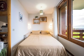 A bedroom with a bed, a window, a shelf with decorative items, and a wall-mounted light. - Le Montagnard - Vue Mont Blanc - Pistes (Landry)