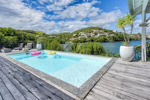A rectangular swimming pool with a wooden deck, surrounded by potted plants and a view of a hillside with houses. - Villa Corail - Piscine et vue mer - Clim (Le Robert)