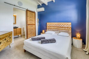 A bedroom with a large bed, bedside lamps, a wooden headboard, and a bathroom with a toilet and sink. - Villa Corail - Piscine et vue mer - Clim (Le Robert)