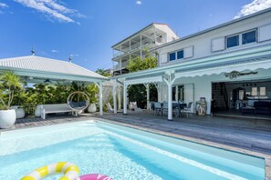 Ein Swimmingpool mit überdachter Terrasse, Sitzgelegenheiten im Freien und Blick auf ein Wohngebäude.