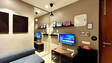A modern hotel room with a sofa, a television, a desk with a computer, and a wall-mounted shelf with framed pictures.