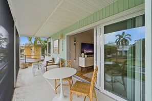 A covered outdoor seating area with a table and chairs, a sliding glass door leading to a room with a painting, and a view of a yellow building and palm trees. - Downtown Naples Walkable Escape – Private Lanai, Steps to Beach & 5th Avenue (Naples)